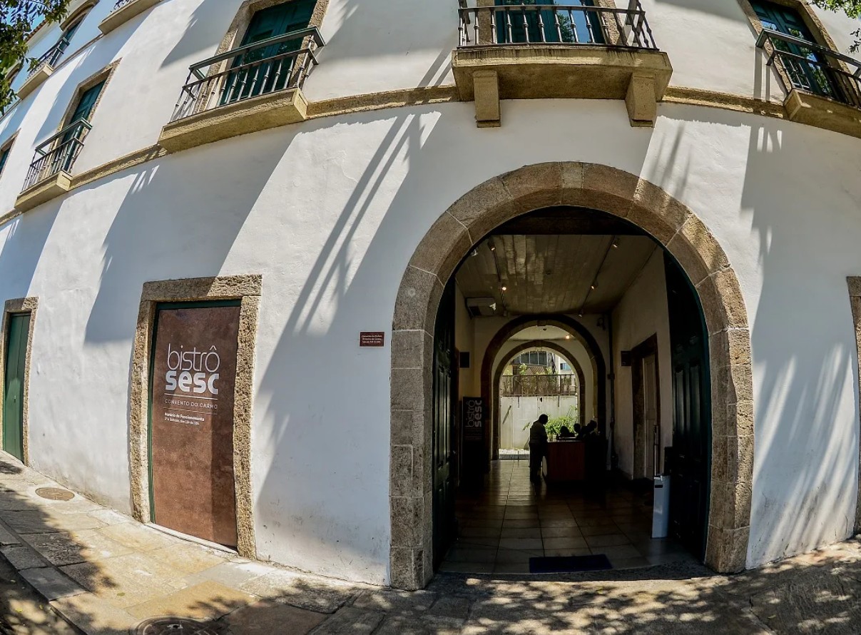 Fachada de um edifício branco com janelas verdes e sacadas de ferro, com uma porta marrom do Bistrô Sesc à esquerda. À direita, um grande arco de pedra leva a um corredor com mais arcos e pessoas ao fundo, sob a luz do sol que projeta sombras no chão e na parede