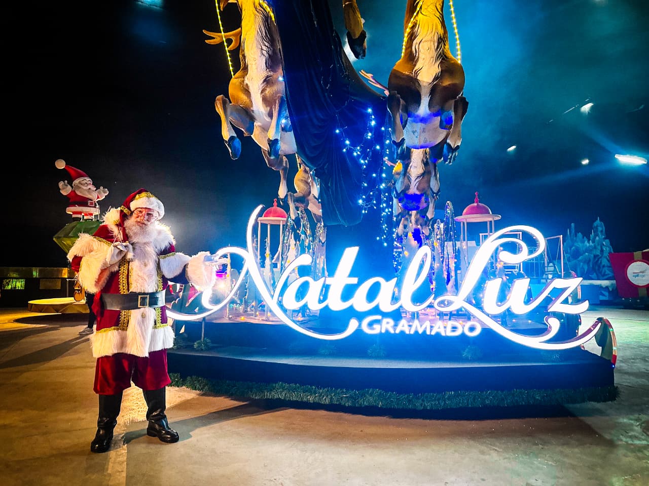 Papai Noel sorridente, com barba branca e roupa vermelha, em frente ao letreiro luminoso Natal Luz Gramado em azul. Ao fundo, um boneco de Papai Noel e decorações natalinas com cavalos e luzes azuis penduradas