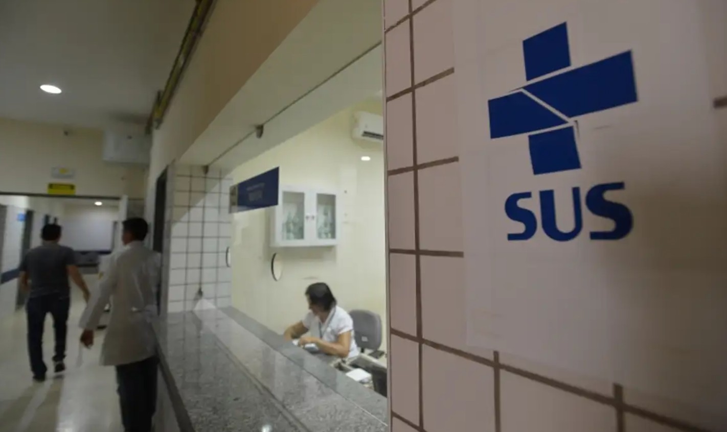 Interior de um hospital ou clínica com o logo azul do SUS em destaque na parede direita. Ao fundo, uma recepcionista atende em um balcão, enquanto dois homens caminham por um corredor iluminado