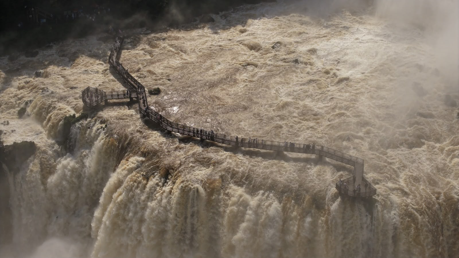 Passarela sinuosa de madeira com muitas pessoas sobre as águas turbulentas e barrentas das Cataratas do Iguaçu, com névoa no ar