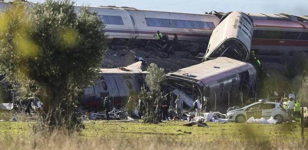 Vagões de trem descarrilados e amassados em um acidente, com equipes de resgate e policiais trabalhando no local. Um carro da Guarda Civil está estacionado na grama, e árvores verdes em primeiro plano em um dia ensolarado