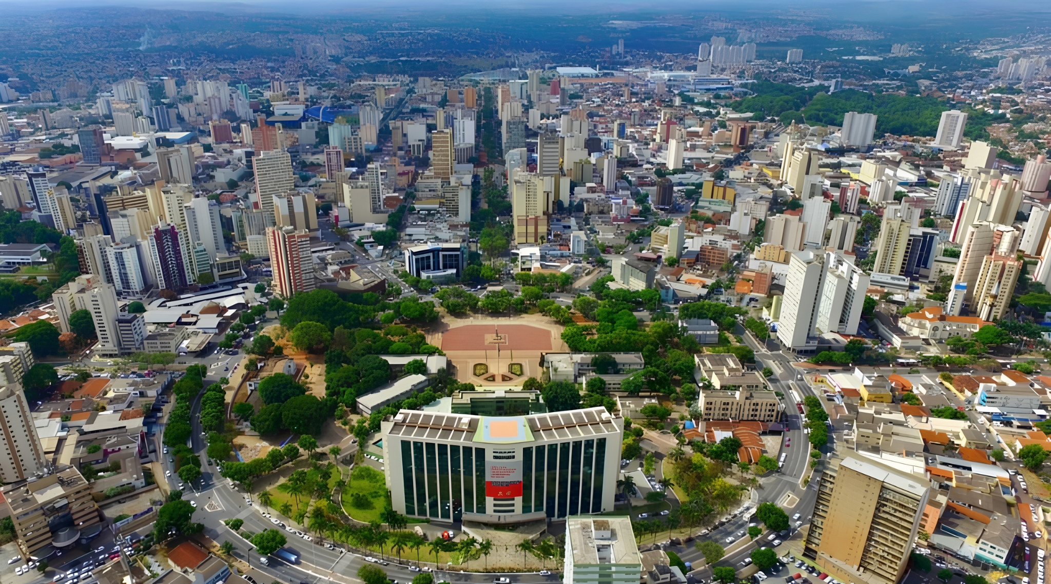 Vista aérea de uma cidade brasileira com muitos prédios altos e árvores verdes, destacando uma praça central e um edifício governamental moderno com telhado solar, cercado por ruas movimentadas e carros estacionados