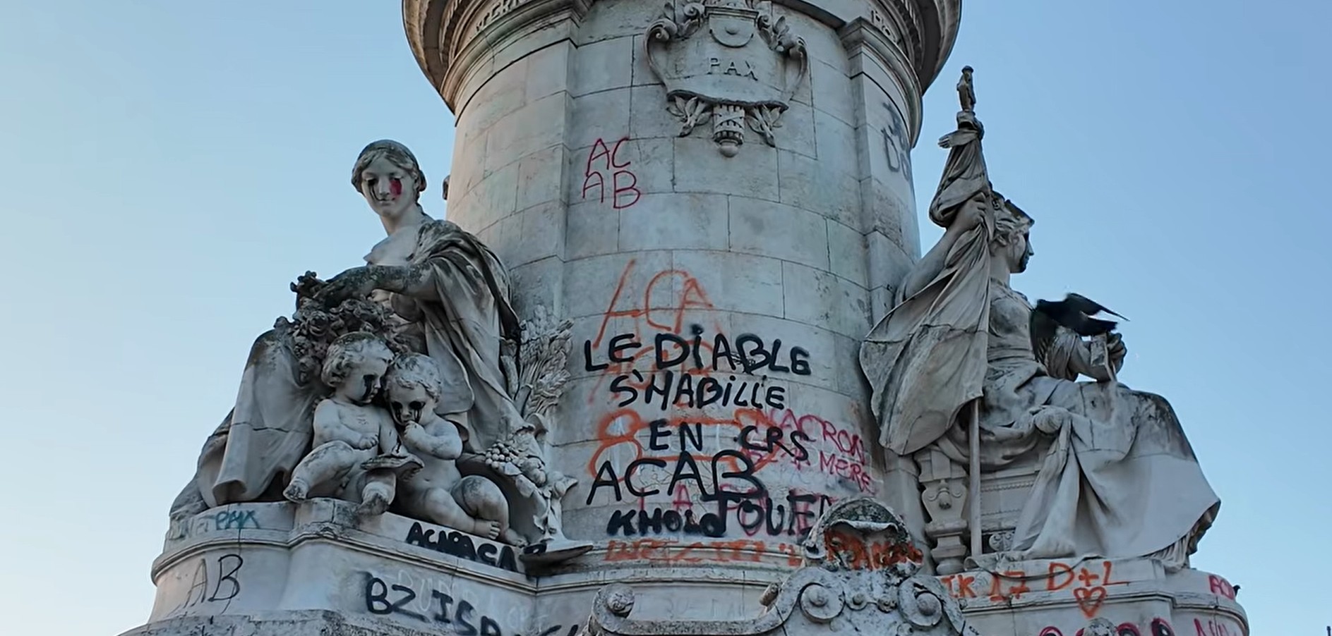 Estátua de pedra com figuras femininas e crianças, coberta por pichações em preto e vermelho, incluindo LE DIABLE SHABILLE EN MACRON e ACAB. Uma das figuras femininas tem tinta vermelha escorrendo do olho, simulando lágrimas de sangue, e as crianças têm tinta preta escorrendo dos olhos. O céu está claro e azul