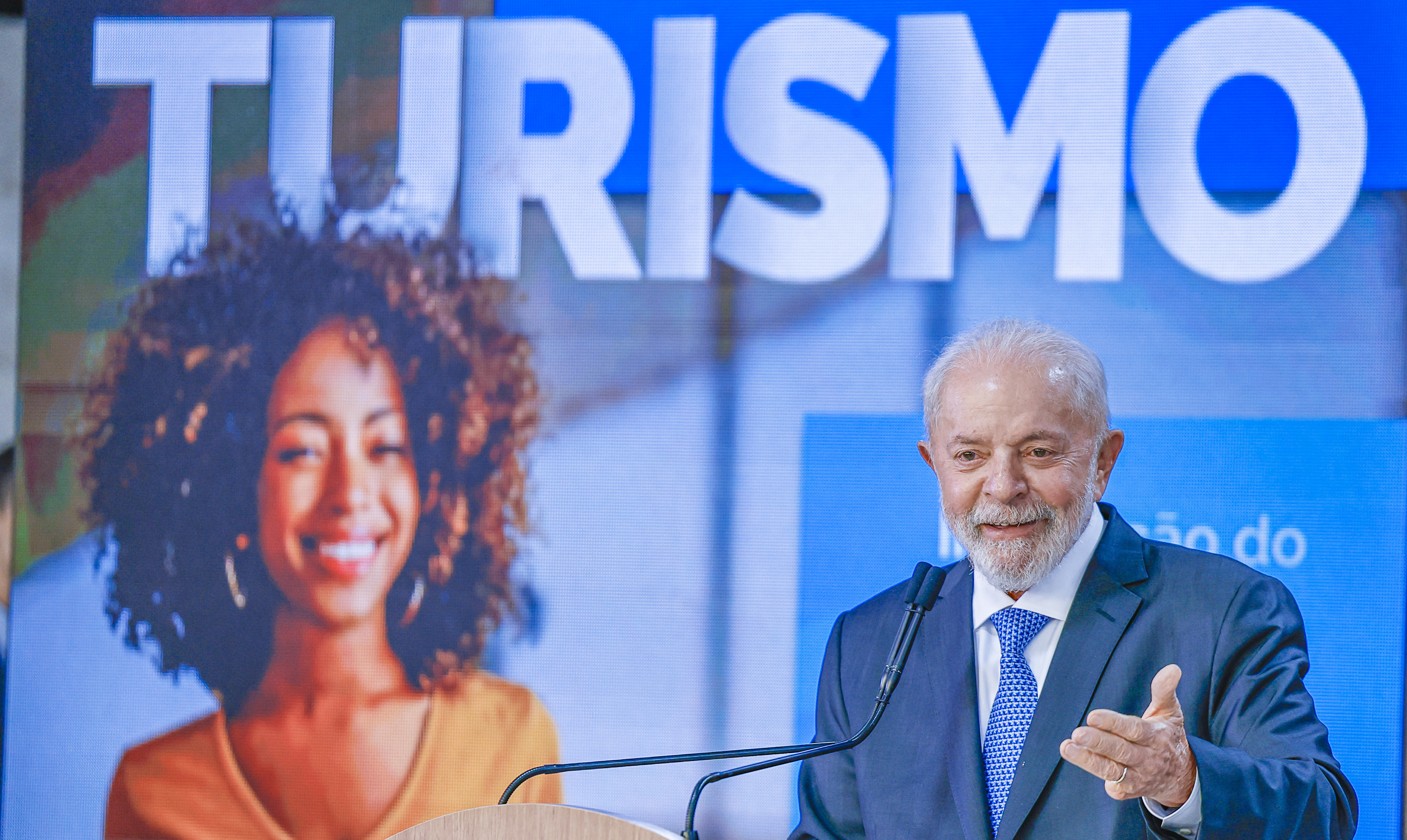 Lula, de terno azul e gravata, sorri e gesticula em um púlpito com microfones. Ao fundo, um telão exibe a palavra TURISMO e a imagem de uma mulher negra sorrindo, com cabelos cacheados.