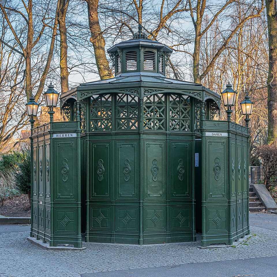 Banheiro público vitoriano verde-escuro, octogonal, com detalhes ornamentados e telhado em cúpula. Placas HERREN e DAMEN indicam as entradas, ladeadas por postes de luz antigos. Árvores sem folhas e céu azul ao fundo, com calçamento de paralelepípedos no primeiro plano