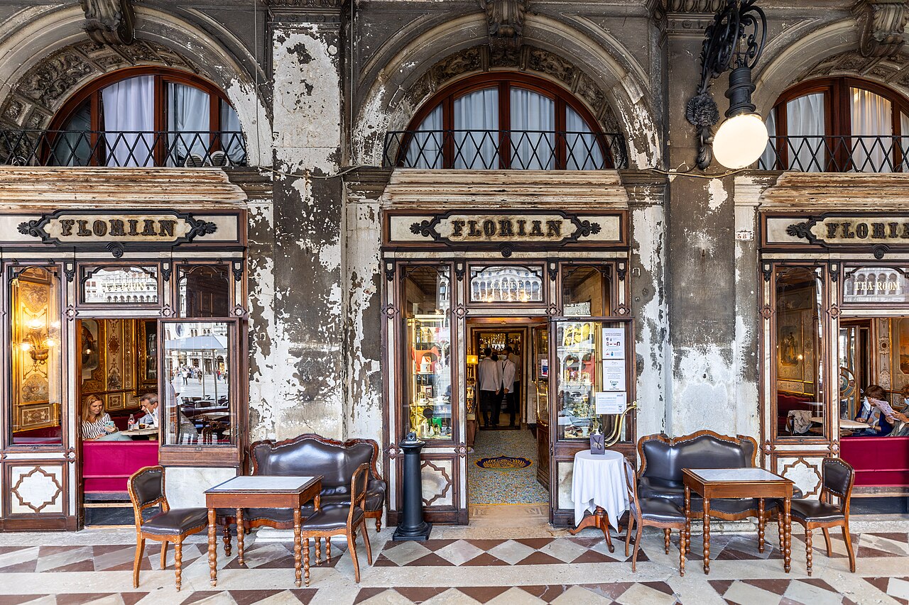 Fachada do Caffè Florian em Veneza, com três arcos superiores e três entradas no térreo. A entrada central, com a placa FLORIAN, tem uma porta aberta e dois funcionários. As entradas laterais têm janelas com pessoas sentadas em sofás vermelhos. Mesas e cadeiras de madeira com estofamento escuro estão dispostas na calçada de piso xadrez, sob a arcada. A parede está descascada, revelando a idade do edifício