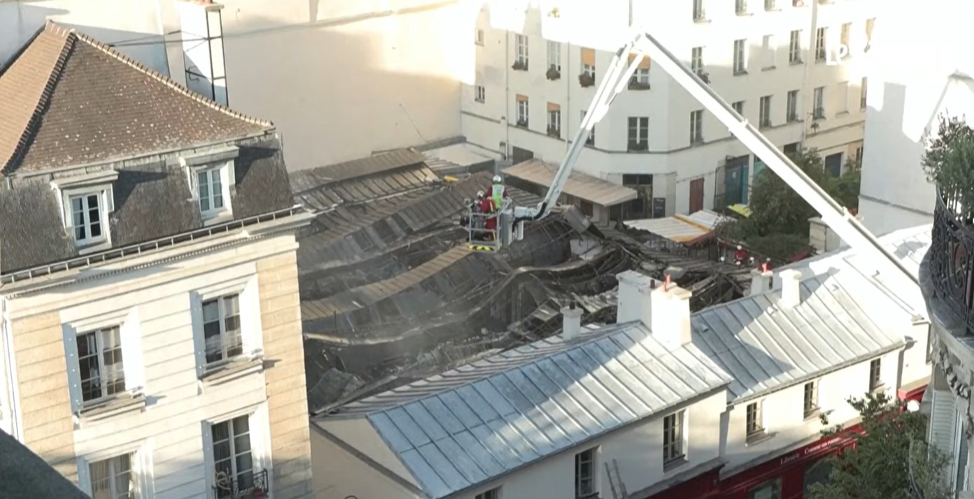 Vista aérea de telhados de edifícios em Paris, com um guindaste branco estendido sobre um telhado colapsado e escuro. Bombeiros em uniformes vermelhos e amarelos estão na cesta do guindaste e no telhado danificado, trabalhando na área afetada. Fumaça branca sobe do telhado destruído, indicando um incêndio recente ou em andamento. Edifícios residenciais de estilo parisiense, com telhados de ardósia e janelas brancas, cercam a cena. O céu está claro e ensolarado